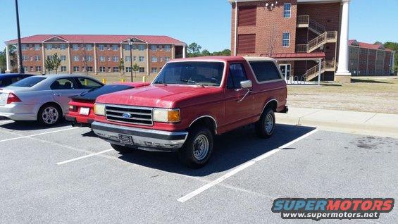 Red Ford Bronco parked in a lot next to other cars, with buildings in the background