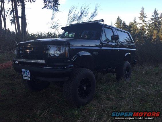 Black 1992 Ford Bronco parked in grassy area with trees behind it. Has a light bar on top
