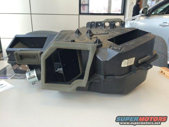HVAC blower motor housing with foam insulation and vents. A silver car is visible in the background