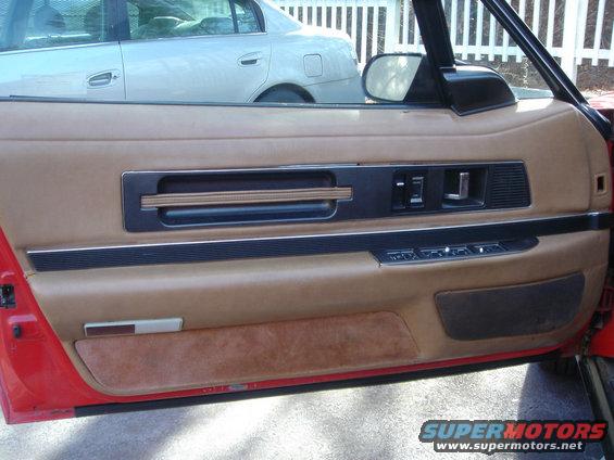 Interior door panel with tan upholstery, black trim, and window controls. Another car is visible through the window
