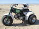 1900 Honda Big Red - A green and white three-wheeled ATV sits on a sandy terrain under a clear sky.