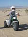 1900 Honda Big Red - A young rider, equipped with a helmet and protective gear, navigates a three-wheeled ATV through a sandy, desert terrain.