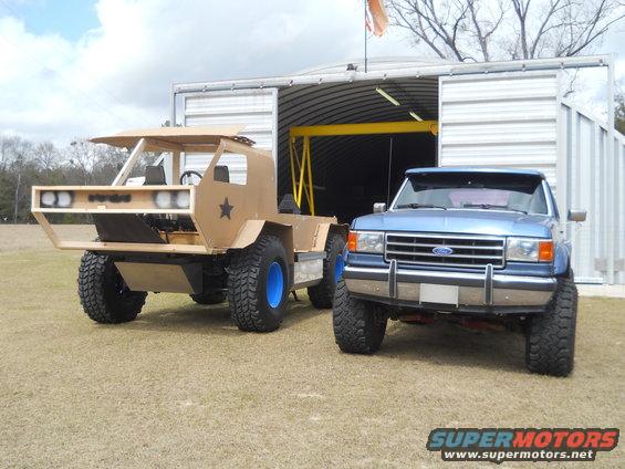 Blue Ford pickup truck parked next to a tan custom-built off-road vehicle in front of a metal building