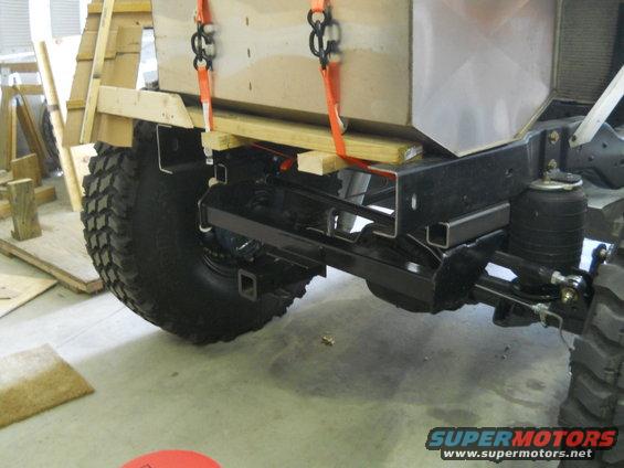 Close-up of a vehicle's rear suspension and frame, with a large wooden crate being lowered by straps