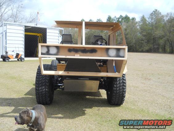 Unfinished wooden vehicle with exposed chassis and large tires, viewed from the rear