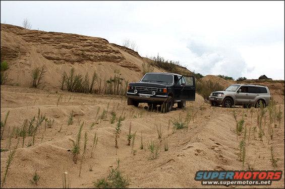 Black SUV with door open and silver SUV on sandy terrain under a cloudy sky