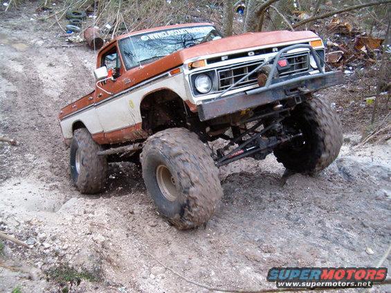 Orange and white Ford F-Series truck with large off-road tires navigating a muddy incline