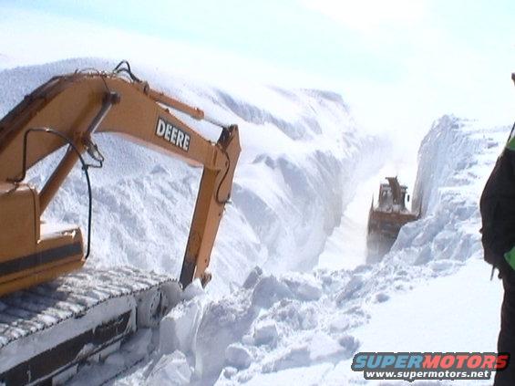 Yellow Deere excavator in foreground, orange bulldozer in background clearing a path through deep snow
