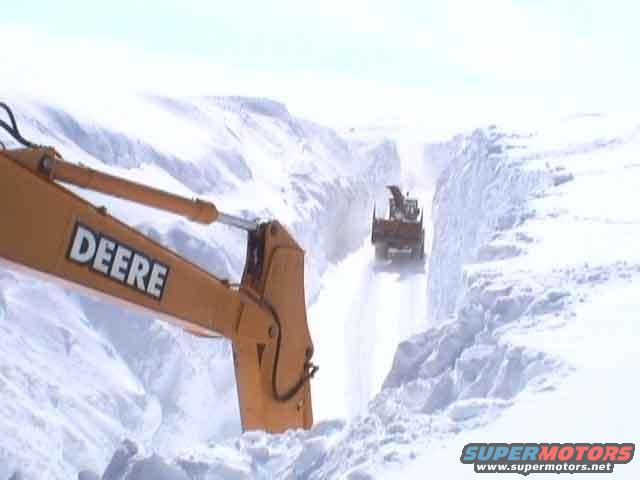 Yellow excavator arm with DEERE logo in foreground, snow blower clearing a path in background