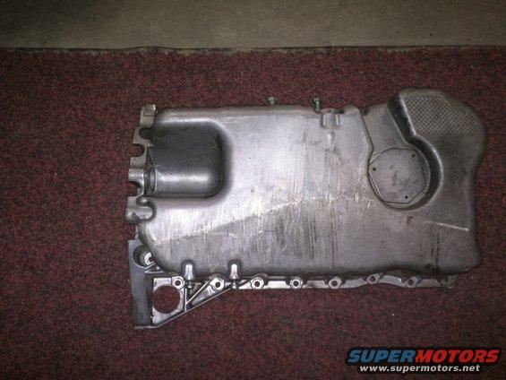 Metal oil pan with mounting holes and a hexagonal cap, resting on a red carpet