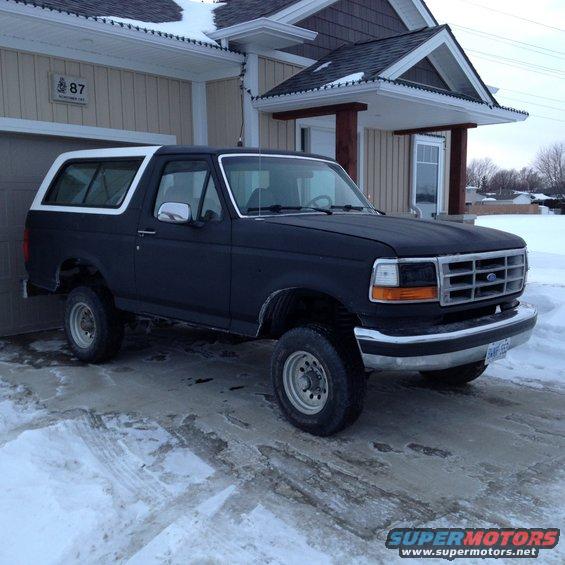 Black Ford Bronco parked in front of a house in the snow. It has a white removable top