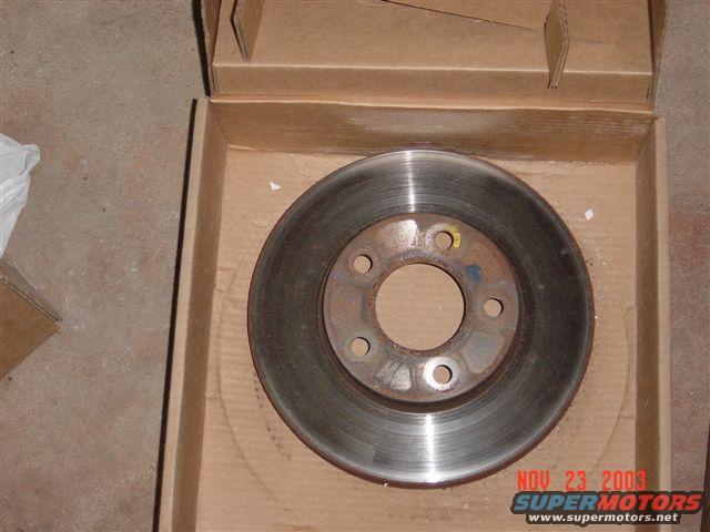 Brake rotor with rust spots, sitting in a cardboard box. Watermark reads NOV 23 2003 SUPERMOTORS.NET