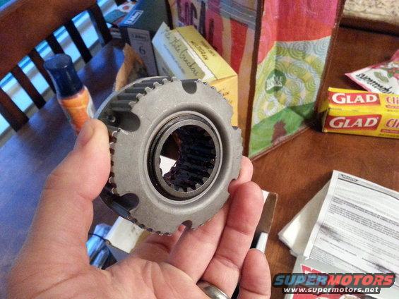 Hand holding a metal hub part with gear teeth and a bearing inside. Boxes and paper are in the background
