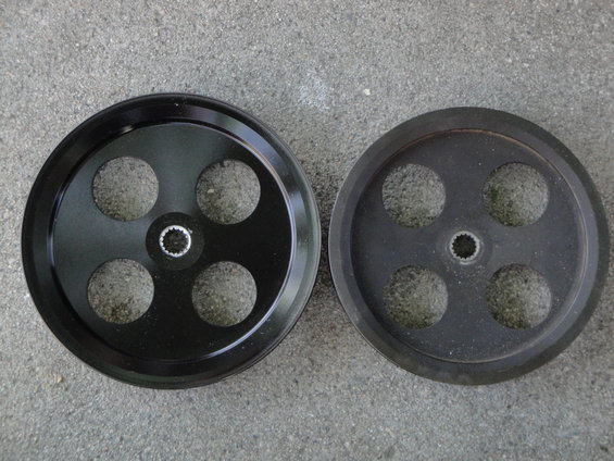 Two black pulleys with four circular cutouts each, one new and one worn, on a concrete surface