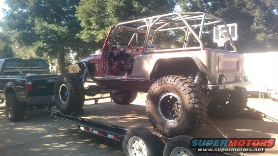 Modified Jeep CJ-series on a trailer, red body, roll cage, large tires, being towed by a black pickup truck