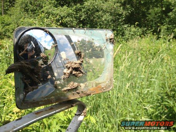 Muddy side mirror on a vehicle, reflecting the sky and a person. A "SUPER MOTORS" logo is visible at the bottom right