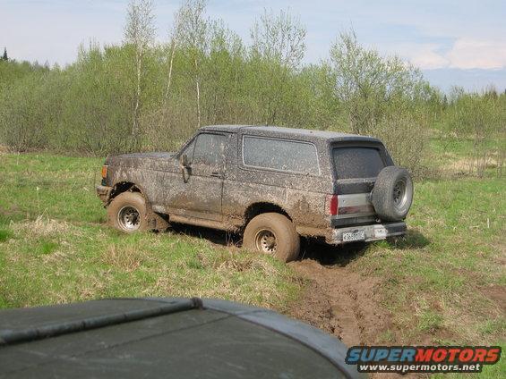 Muddy 1987 Ford Bronco stuck in a muddy field, with another vehicle's hood visible in the foreground