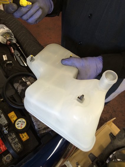 hands in gloves holding a white plastic coolant reservoir with yellow caps on mounting posts