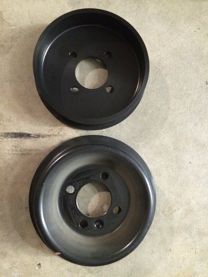Two black automotive pulleys, one new and one used, on a concrete surface