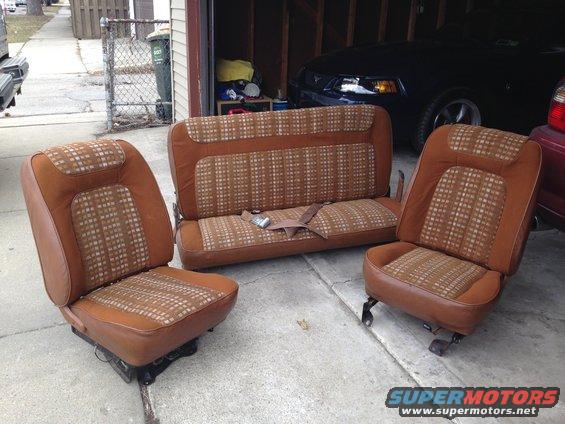 Three brown plaid car seats and a bench seat are on a concrete surface in front of a garage. A dark car is parked in the garage