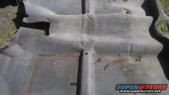 Worn gray car carpet with rust stains and visible floor pan indentations
