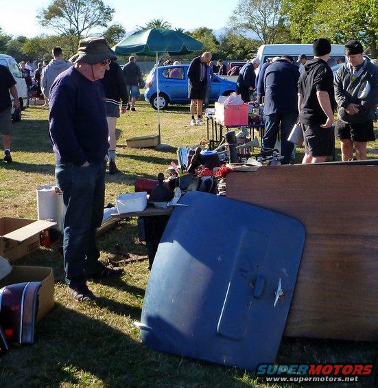 People browsing car parts and other items at an outdoor market. A blue hood is in the foreground