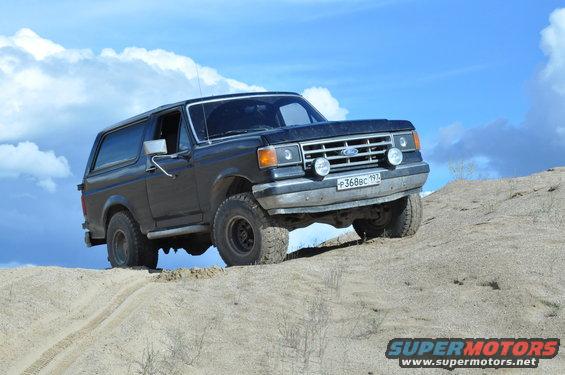 Black 1987 Ford Bronco driving up a sandy hill under a blue sky with clouds