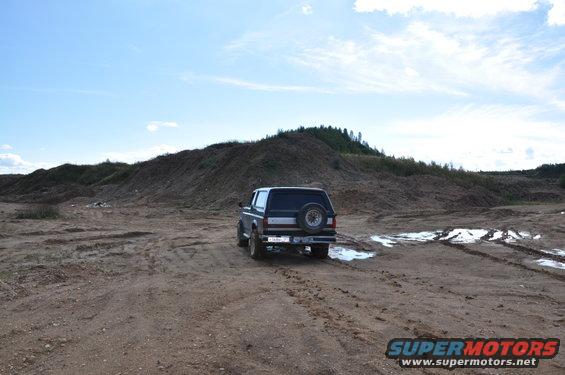 Dark blue Ford Bronco parked on dirt road with tire tracks and puddles. Hill in background