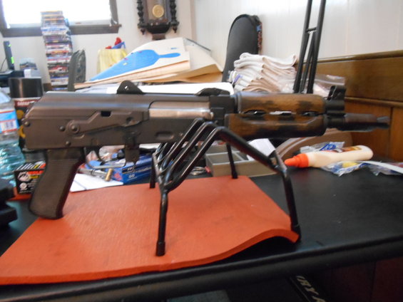 AK-pattern rifle on a black metal stand, resting on an orange mat on a desk