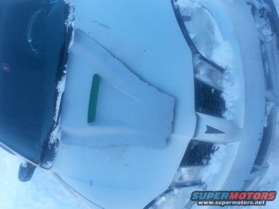 White car hood covered in snow, with a green vent and a black grille visible