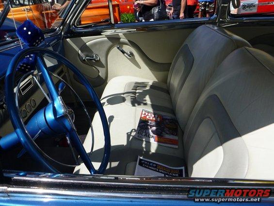 Interior view of a classic car with a blue steering wheel, fuzzy dice, and a magazine on the seat