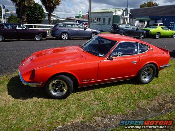 Red Datsun 240Z parked on grass, other cars and buildings in background