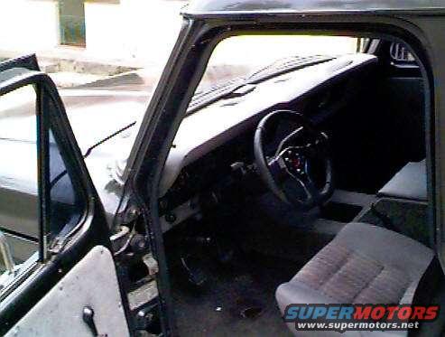 Interior of a dark-colored Ford F-100 truck, showing the driver's seat, dashboard, and steering wheel. Watermark "SUPERMOTORS www.supermotors.net" is visible