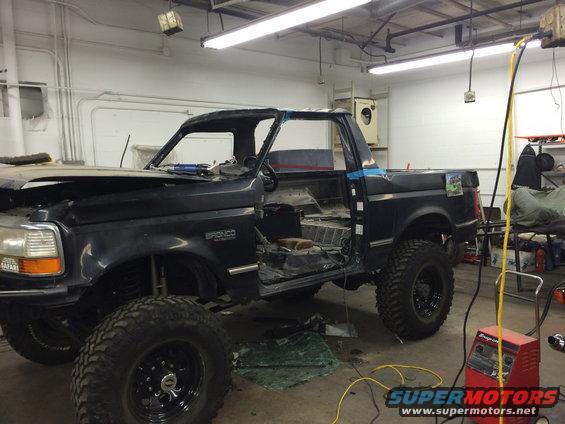 Black Ford Bronco with roof removed, doors open, in a workshop. It has large off-road tires and appears to be undergoing modifications