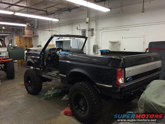 Dark blue Ford Bronco with roof removed, lifted on large tires, in a garage. Another vehicle is partially visible to the left