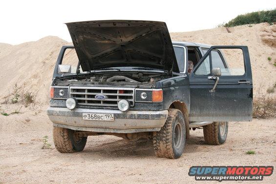 Front view of a dark 1987 Ford Bronco parked on dirt, hood and driver's door open, showing the engine