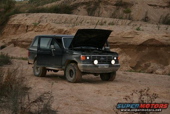 Black Ford Bronco with hood open, parked on a dirt road with sandy hills in the background