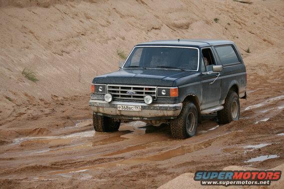1987 Ford Bronco driving on a muddy track with sand dunes in background