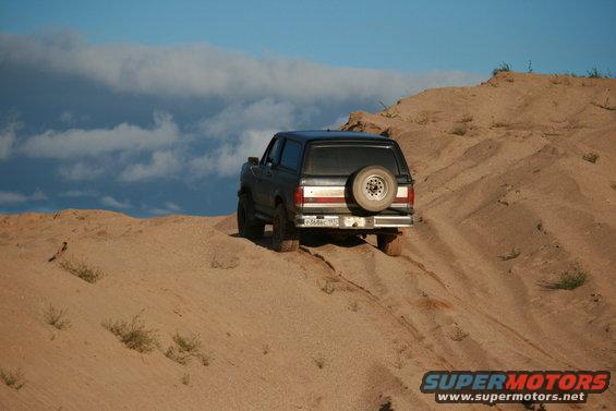 Black 1987 Ford Bronco driving up a sand dune with a spare tire on the back. Blue sky with clouds above