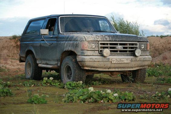 1987 Ford Bronco parked on dirt with vegetation, covered in mud