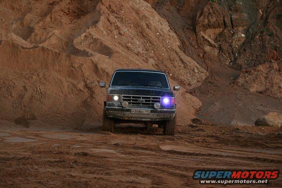 Front view of a dark-colored 1987 Ford Bronco driving on a dirt road with sandy hills in the background