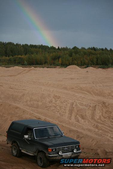 Dark SUV parked on sand dunes with a rainbow in the sky