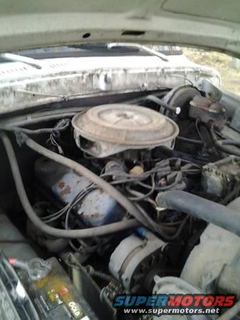 Engine bay view of an older vehicle, showing the carburetor, alternator, and battery