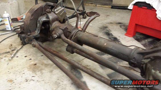 Rusty front axle assembly with steering linkage and brake components, lying on a concrete floor
