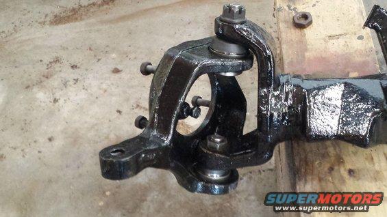 Close-up of a black metal vehicle suspension component, possibly a spindle or knuckle, with bolts and a ball joint visible