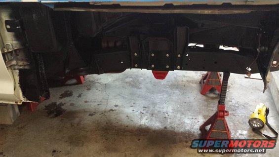 Underside of a vehicle on jack stands, showing the frame and suspension components. A yellow work light is on the floor