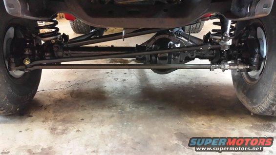 Underside view of front axle and suspension components on a vehicle. Another vehicle is visible in the background