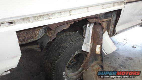 White vehicle's rear wheel well showing extensive rust and damage. Some metal patches are visible