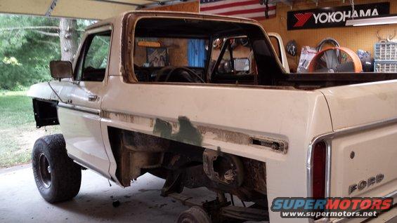 White Ford Bronco, missing doors and fenders, in a garage. The word "FORD" is visible on the tailgate
