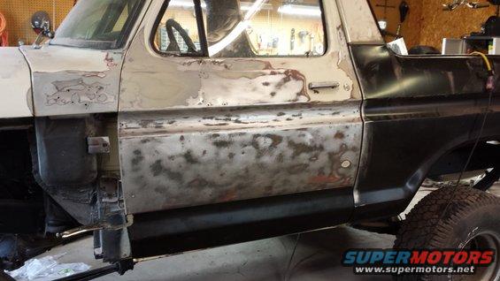 Side view of a Ford Bronco with primer and bare metal body panels, showing rust spots and bodywork in progress